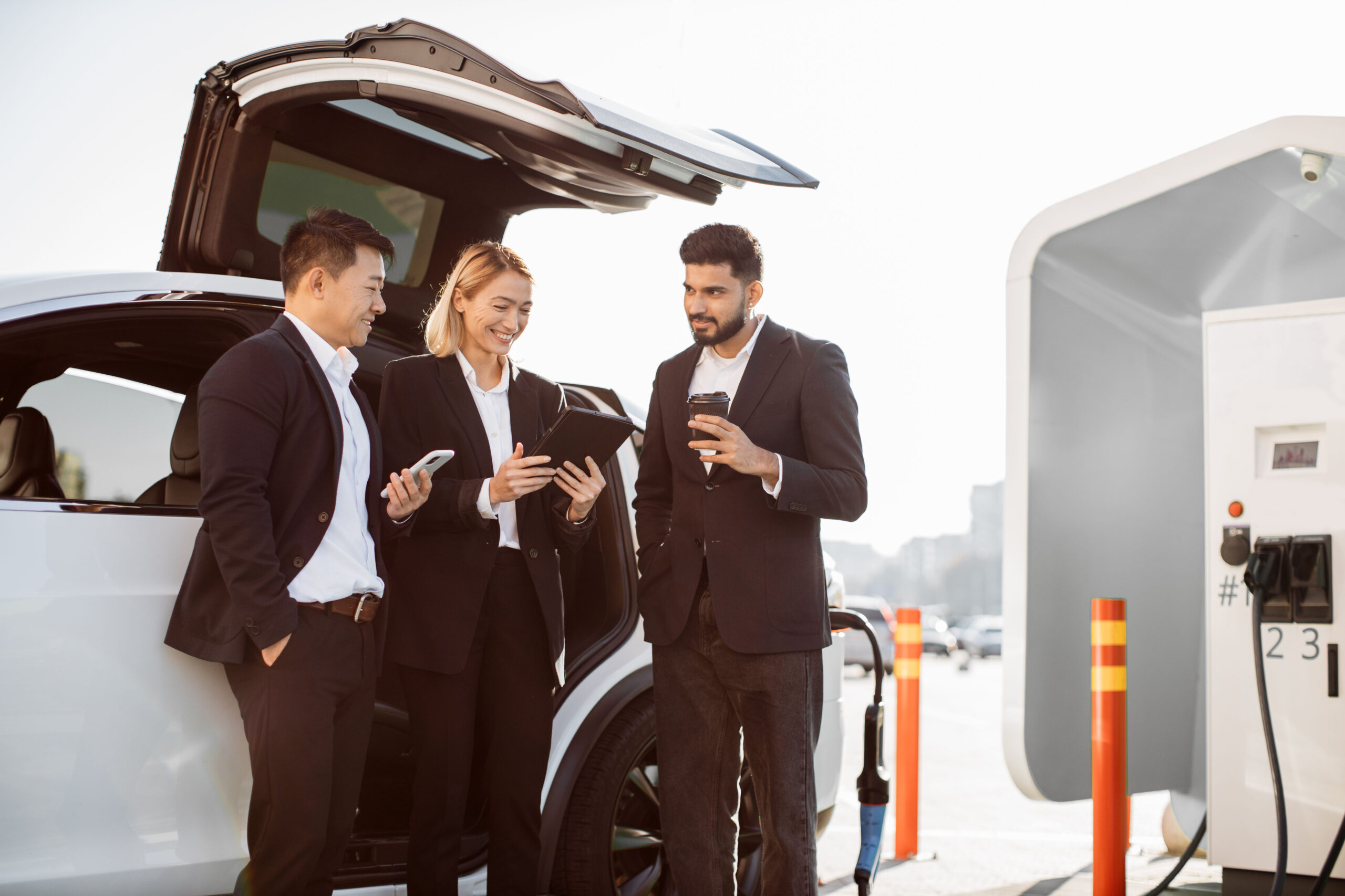 Multiethnic business people studying project on tablet while charging electric car at station. Arabic man, caucasian woman and asian male in formal outfit using waiting time for coffee and work.